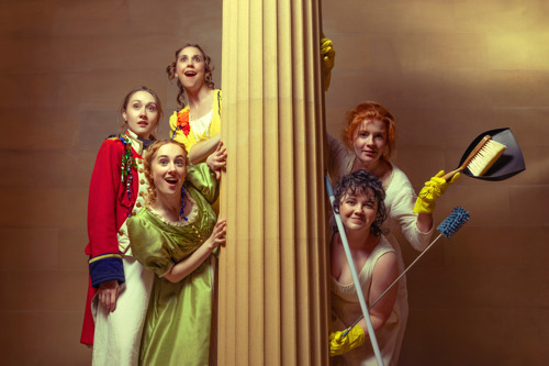 5 women peeping from behind a pillar - the 3 on the left are in formal period dress and the two on the right are in rubber gloves holding cleaning equipment